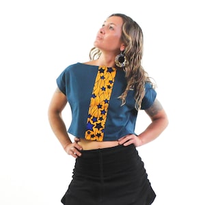 K&ouml;nnte beinhalten: Eine Frau tr&auml;gt ein blaues Crop-Top mit einem gelben und blauen Muster. Das Top hat kurze &Auml;rmel und eine lockere Passform. Sie tr&auml;gt auch einen schwarzen Minirock.