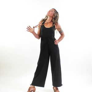May include: A woman wearing a black jumpsuit with a beaded necklace. She is standing with her head tilted back and her arms outstretched.