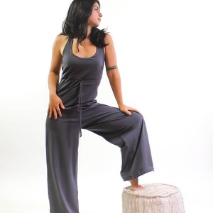May include: A woman wearing a gray jumpsuit with a drawstring waist stands with one foot on a white and gray pouf.