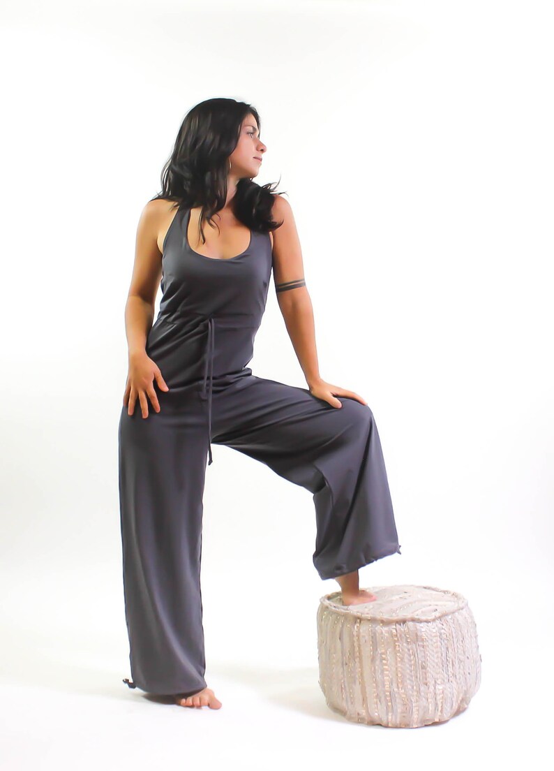 May include: A woman wearing a gray jumpsuit with a drawstring waist stands with one foot on a white and gray pouf.