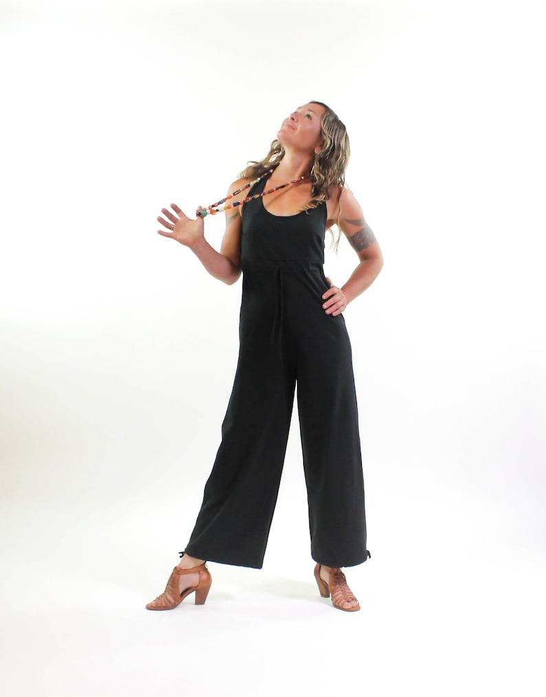 May include: A woman wearing a black jumpsuit with a beaded necklace. She is standing with her head tilted back and her arms outstretched.