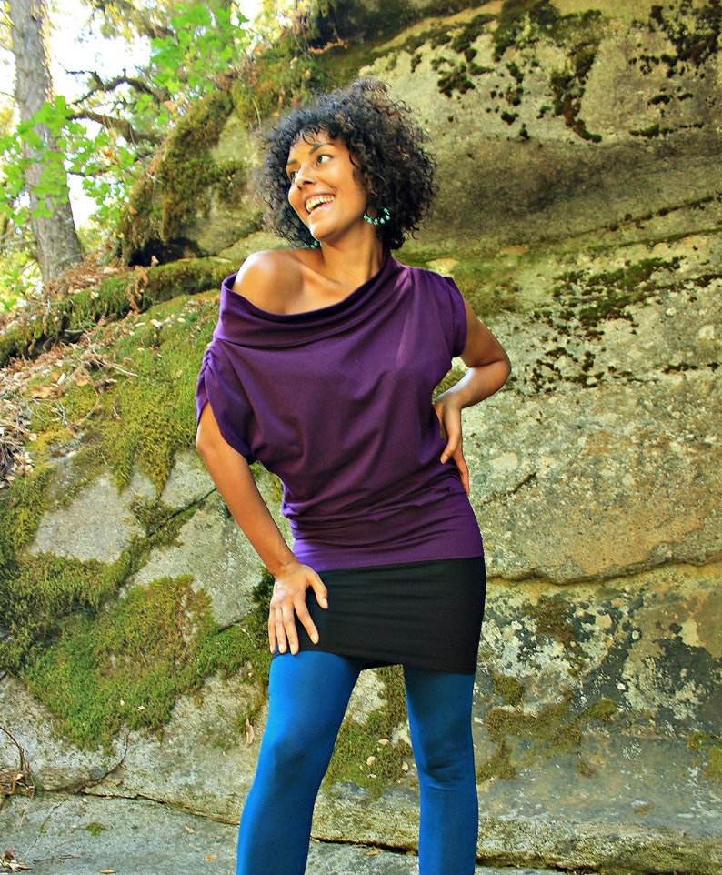 May include: A woman wearing a purple off-the-shoulder top with a black skirt and blue leggings. She is standing in front of a mossy rock.