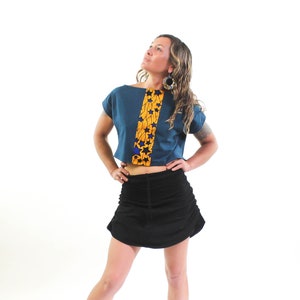 Puede incluir: Una mujer con un top corto azul turquesa con un panel estampado en amarillo y azul. El top tiene mangas cortas y cuello barco. Tambi&eacute;n lleva una falda negra.