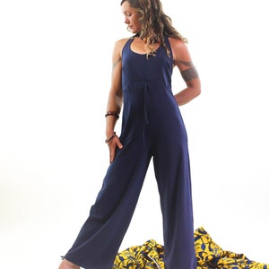 May include: A woman wearing a navy blue jumpsuit with a halter neckline and a tie at the waist. She is standing on a yellow and blue patterned fabric.