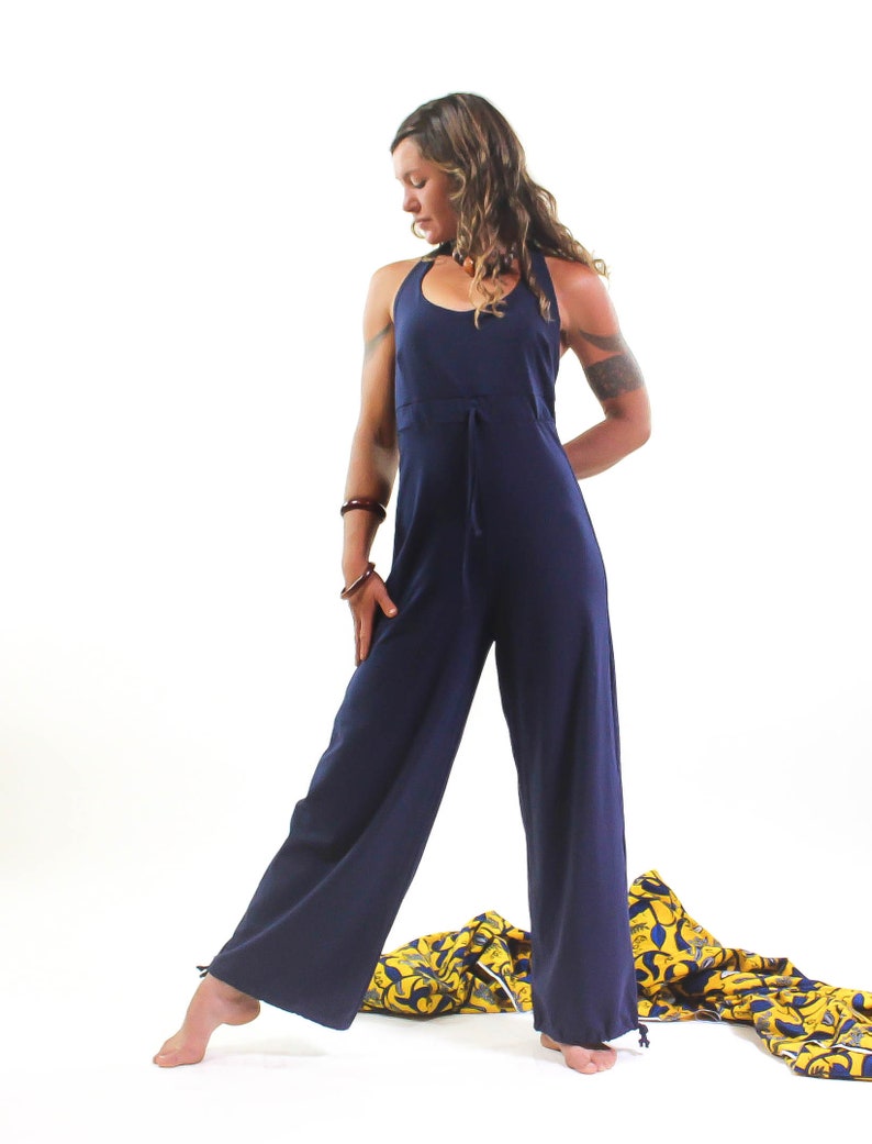 May include: A woman wearing a navy blue jumpsuit with a halter neckline and a tie at the waist. She is standing on a yellow and blue patterned fabric.