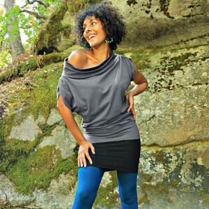 Op de afbeelding: Een vrouw draagt een grijs, off-shoulder top met een zwarte rok en blauwe panty's. Ze staat voor een mossige rots.