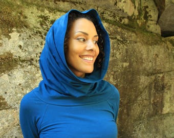 May include: A woman wearing a blue cowl neck sweater with a hood. She is standing in front of a rocky background.