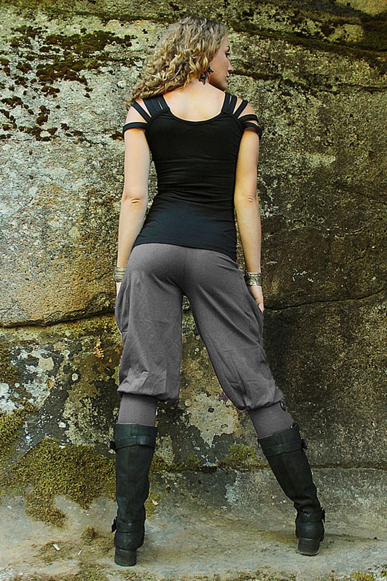 May include: A black top with strap details and gray capri pants are paired with black boots. The outfit is set against a textured stone wall, creating a contrast between the soft clothing and the rough background.