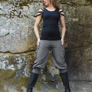 May include: A woman wearing a black tank top with cut-out shoulders and gray cropped pants. She is standing in front of a rocky wall with moss growing on it. She is wearing black boots.
