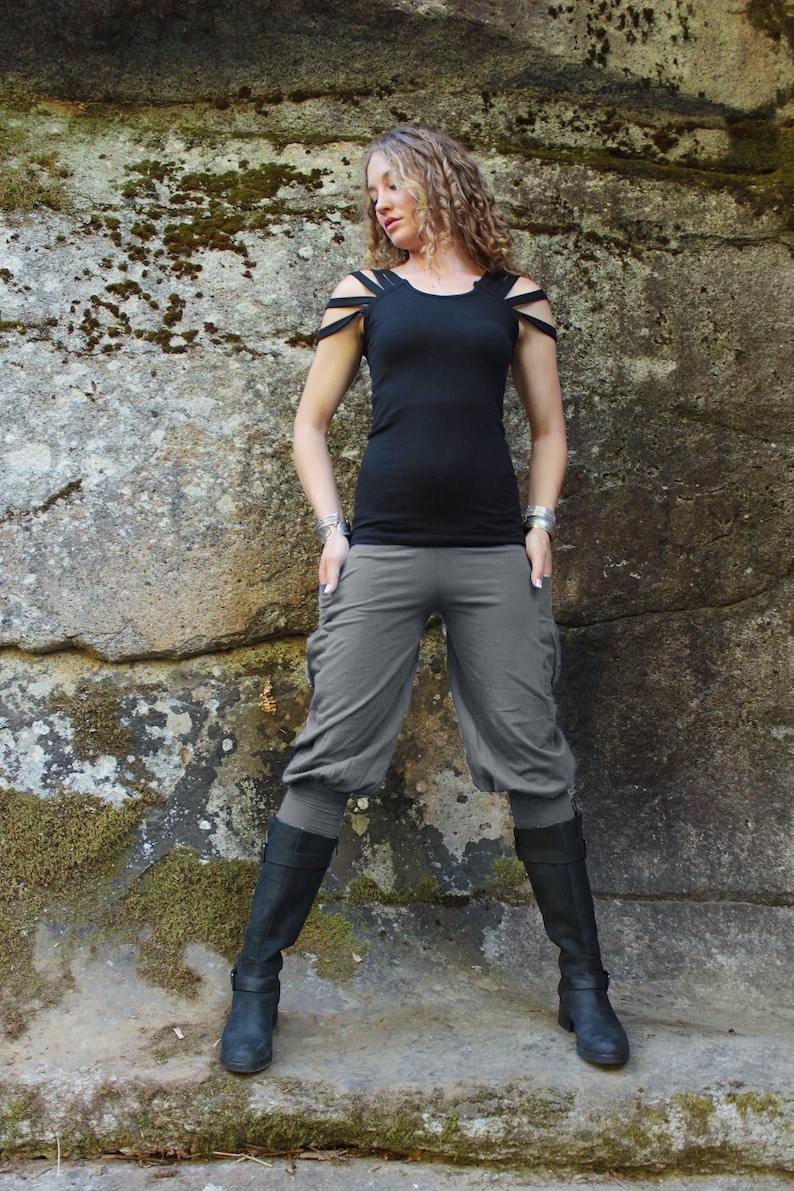 May include: A woman wearing a black tank top with cut-out shoulders and gray cropped pants. She is standing in front of a rocky wall with moss growing on it. She is wearing black boots.