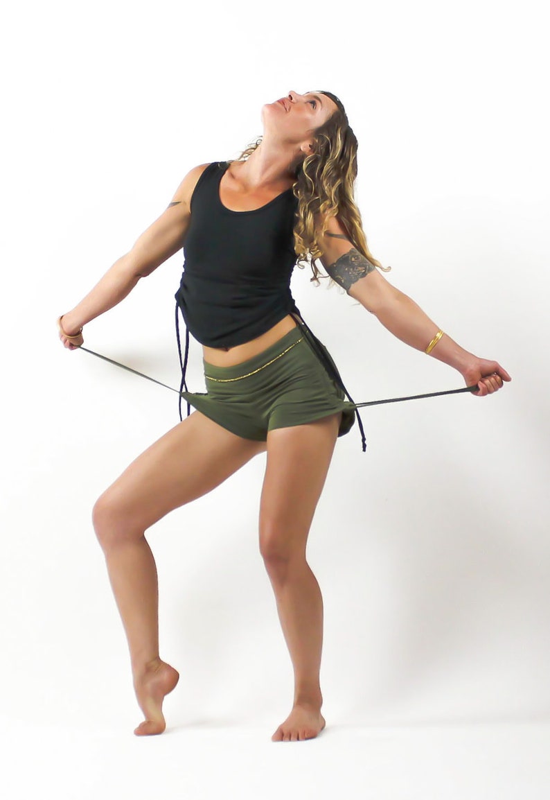 Op de afbeelding: Een vrouw draagt een zwart tanktop en olijfgroene shorts met een gouden kettingband. Ze houdt een zwart koord in haar handen en kijkt omhoog.