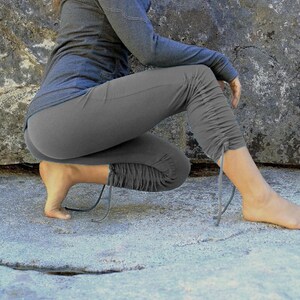 May include: A person wearing gray yoga pants with a drawstring detail at the ankle. The pants are gathered at the knee and the person is sitting on a large rock.