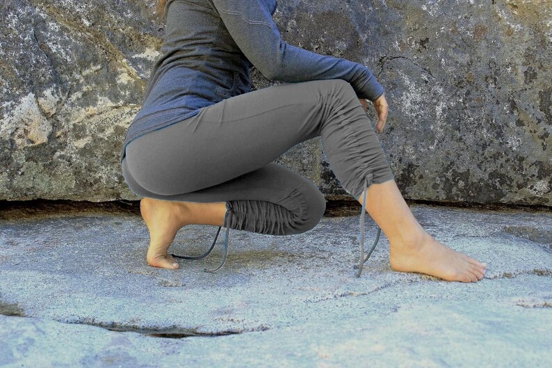 May include: A person wearing gray yoga pants with a drawstring detail at the ankle. The pants are gathered at the knee and the person is sitting on a large rock.