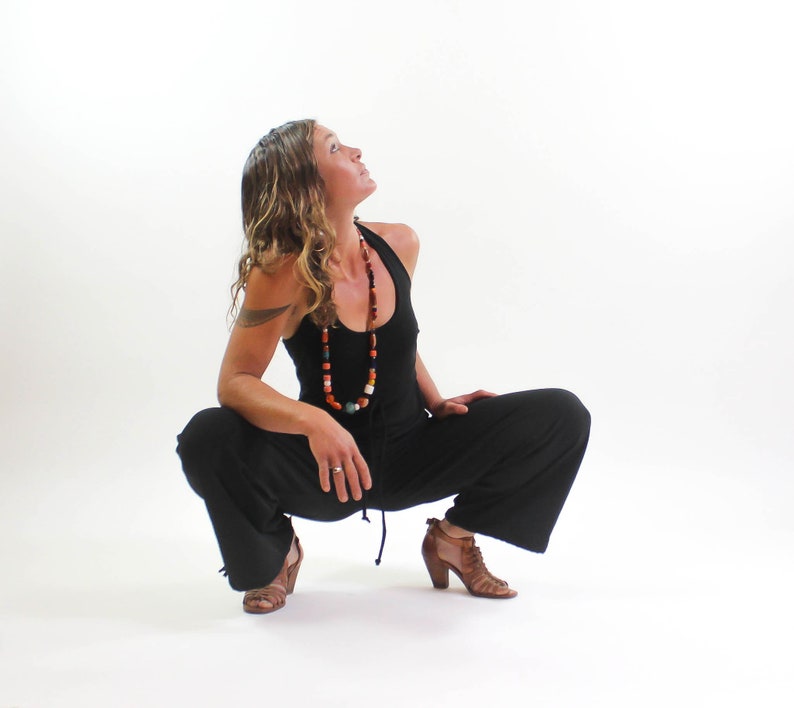 May include: A woman wearing a black jumpsuit with a colorful beaded necklace, sitting with her legs crossed and looking up.