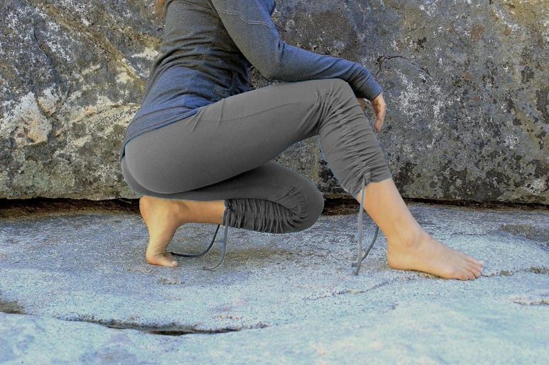 May include: Gray athletic pants with drawstring details at the ankles. The pants are paired with a long-sleeved top. The person is squatting on a light gray stone surface, with a large rock in the background. The outfit appears to be for outdoor activities.