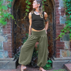 May include: A woman wearing a black tank top and olive green, wide-legged pants. She is standing in front of a brick wall with ivy growing on it.