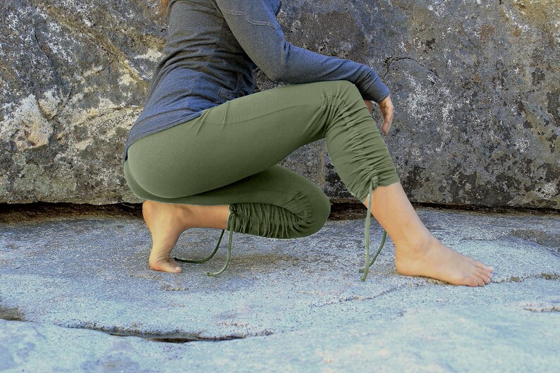 May include: Olive green cropped leggings with drawstring details at the ankles. The leggings are paired with a long-sleeved gray top. The person is squatting on a light gray stone surface, with a large rock in the background.
