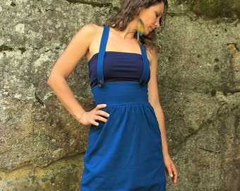 May include: A woman wearing a blue denim overall dress with a dark blue top underneath. The dress has a high waist and a gathered skirt. She is standing in front of a rocky background.