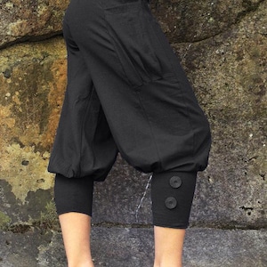 May include: Black cropped pants with a loose, gathered fit and a button detail at the bottom of the legs. The pants have a large pocket on the left side.