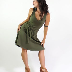 May include: A woman wearing a green sleeveless dress with a tie at the waist. She is standing with her right leg bent and her left leg straight. She is wearing brown sandals.