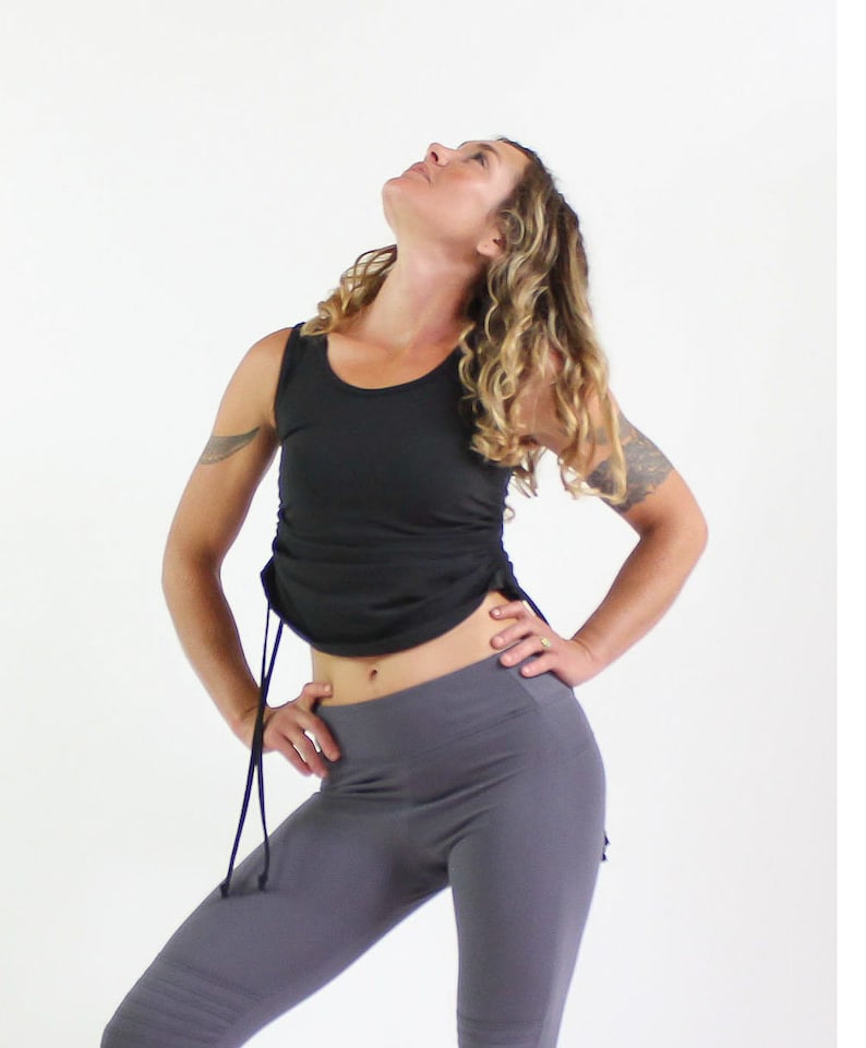 May include: A woman wearing a black tank top with a drawstring detail and gray leggings. The tank top has a scoop neck and the leggings are fitted.