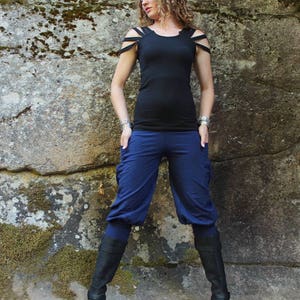 May include: A woman wearing a black top with cut-out shoulder straps and navy blue cropped pants. She is also wearing black boots. The background is a large stone wall.
