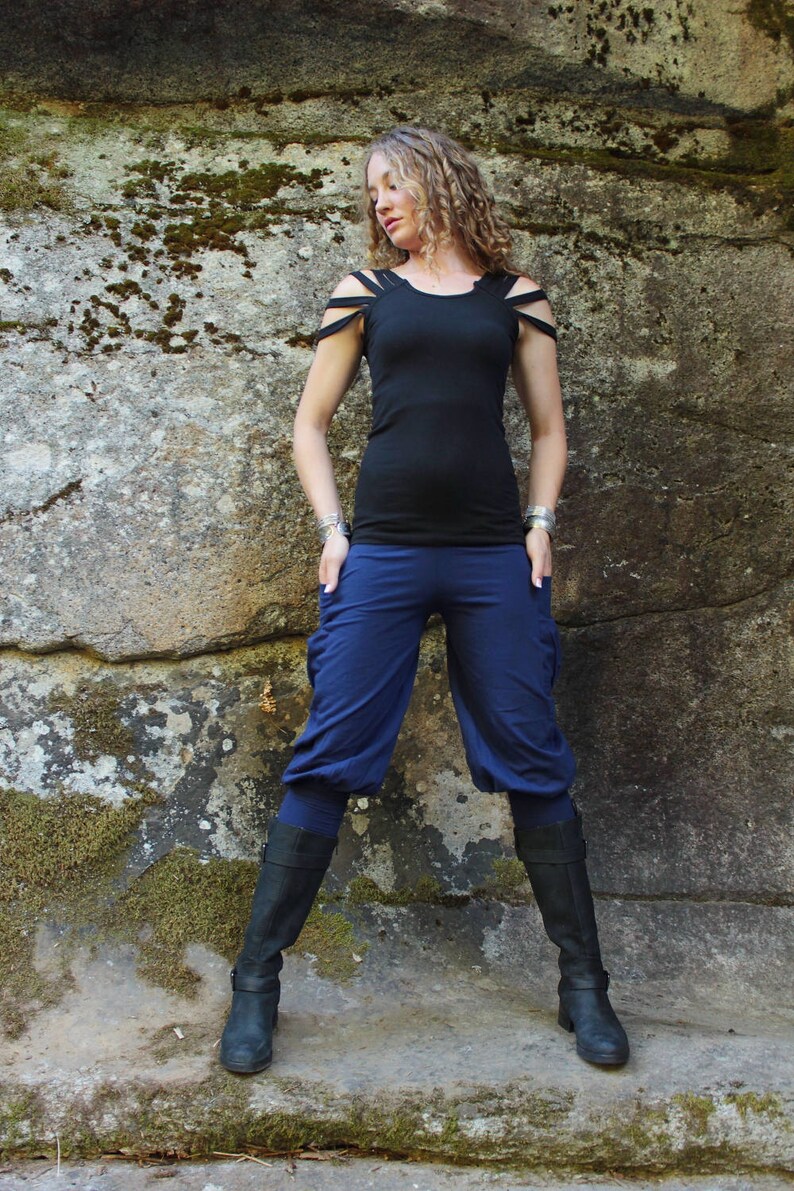 May include: A woman wearing a black top with cut-out shoulder straps and navy blue cropped pants. She is also wearing black boots. The background is a large stone wall.