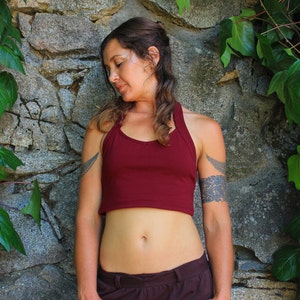 May include: A woman wearing a maroon crop top with a halter neckline. She has a tattoo on her upper arm and is standing in front of a stone wall with ivy growing on it.