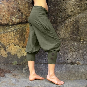 May include: Olive green cropped trousers with a relaxed fit. The trousers feature a gathered ankle with two decorative buttons. The fabric appears to be a soft, breathable material. The trousers have a pocket on the side.