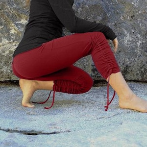 Puede incluir: Una persona con una parte superior negra de manga larga y pantalones cortos de color burdeos con detalles de cordón en los tobillos. La persona está en cuclillas sobre una superficie de piedra gris claro, con una gran roca en el fondo.