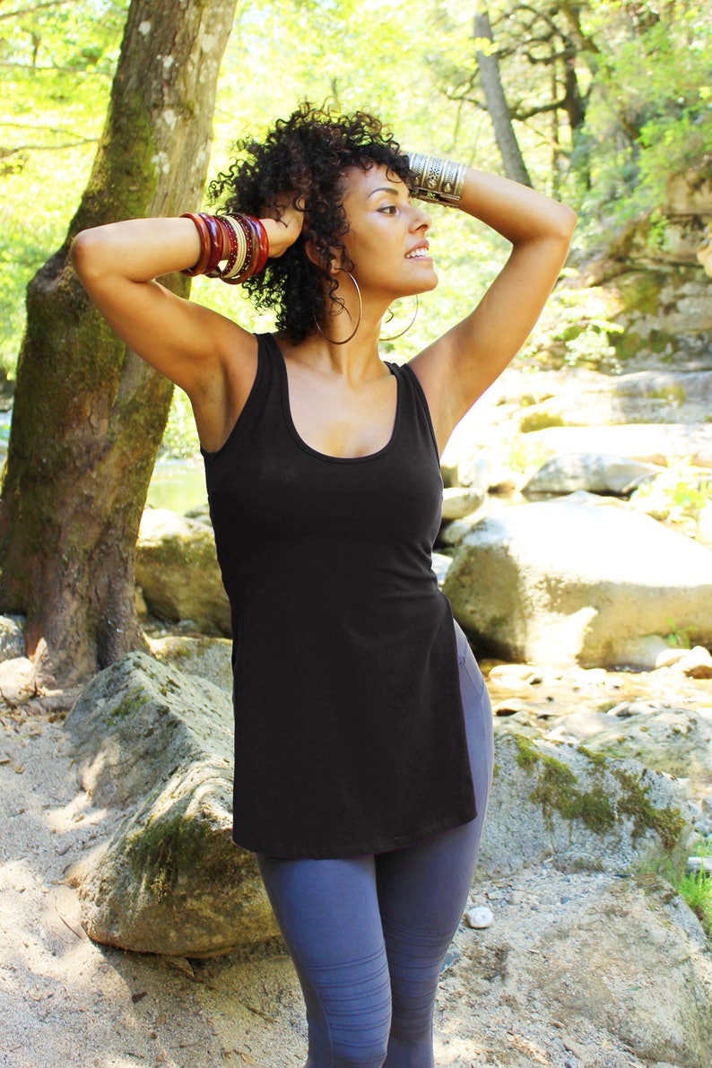 May include: A woman with curly dark hair wearing a black tank top with a side slit and dark blue leggings. She is wearing multiple bracelets on her wrist and a large hoop earring.