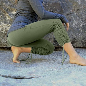 May include: A person wearing olive green yoga pants with a drawstring detail at the ankles. The pants are gathered at the knees and the person is sitting on a rock.