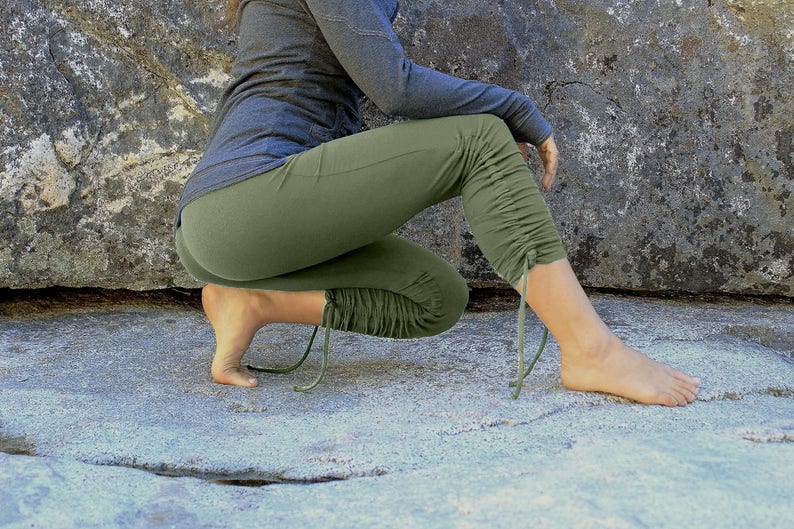 May include: A person wearing olive green yoga pants with a drawstring detail at the ankles. The pants are gathered at the knees and the person is sitting on a rock.