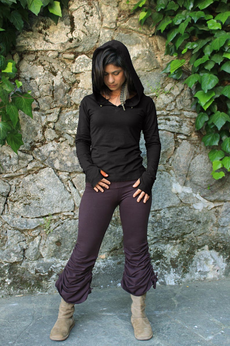 May include: A woman wearing a black hooded top with a long sleeve and a dark purple pair of pants with a gathered detail at the bottom. She is standing in front of a stone wall with green ivy growing on it.