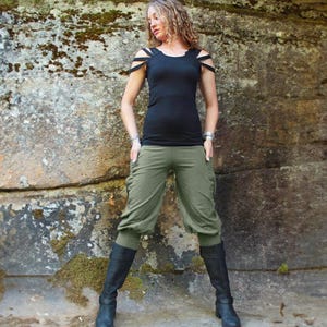 May include: A woman wearing a black top with cut-out shoulder straps, olive green capri pants, and black boots. The outfit is set against a textured stone background, creating a contrast between the clothing and the natural setting.