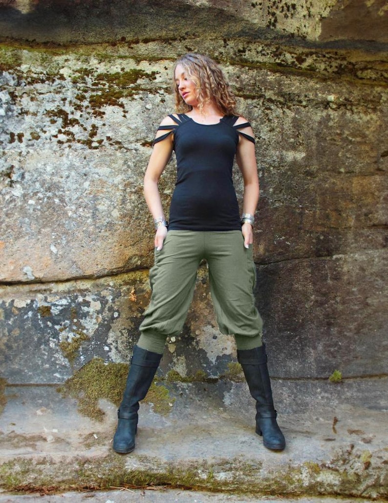 May include: A woman wearing a black top with cut-out shoulder straps, olive green capri pants, and black boots. The outfit is set against a textured stone background, creating a contrast between the clothing and the natural setting.