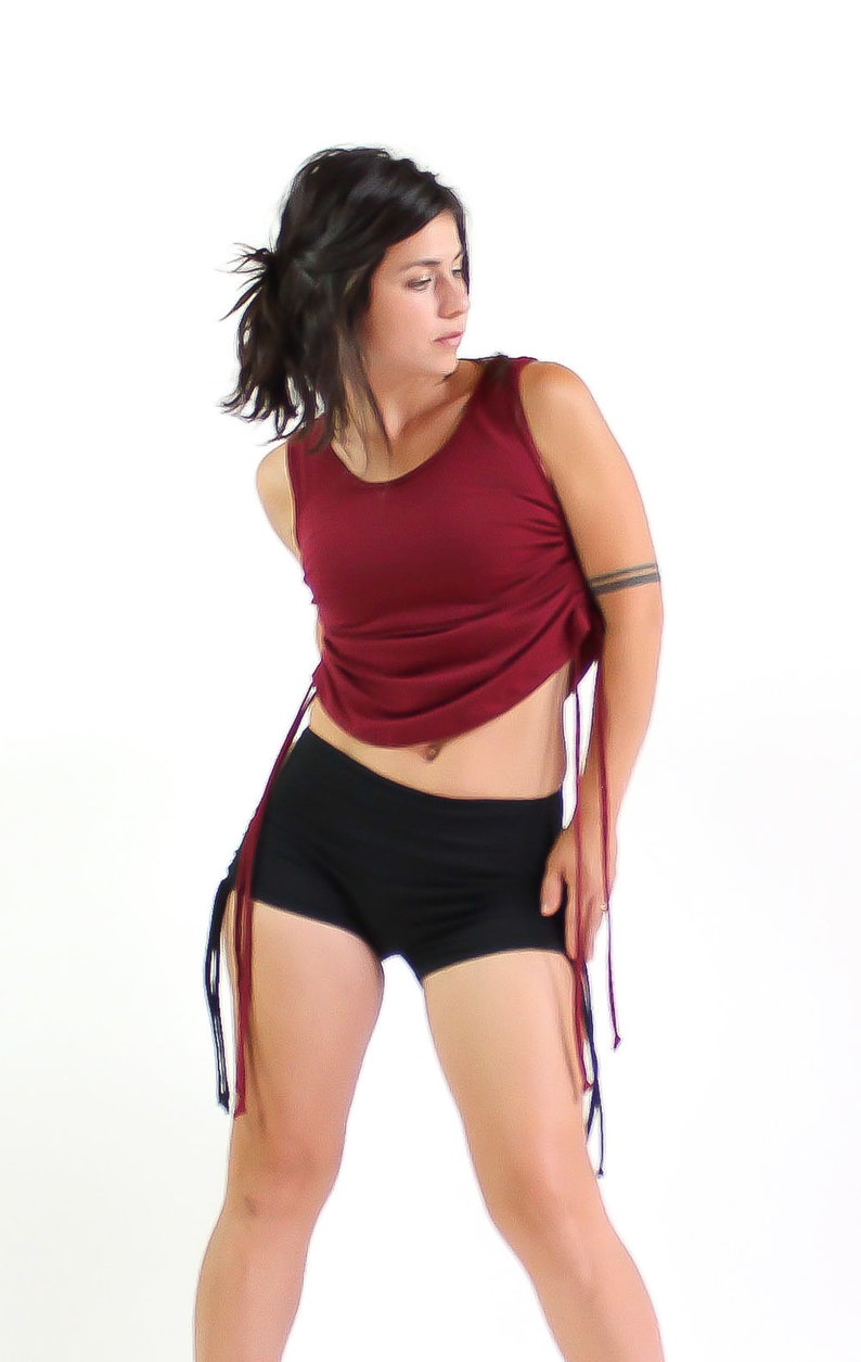 May include: A woman wearing a maroon tank top with side ties and black shorts. The tank top is cropped and the shorts are high-waisted.