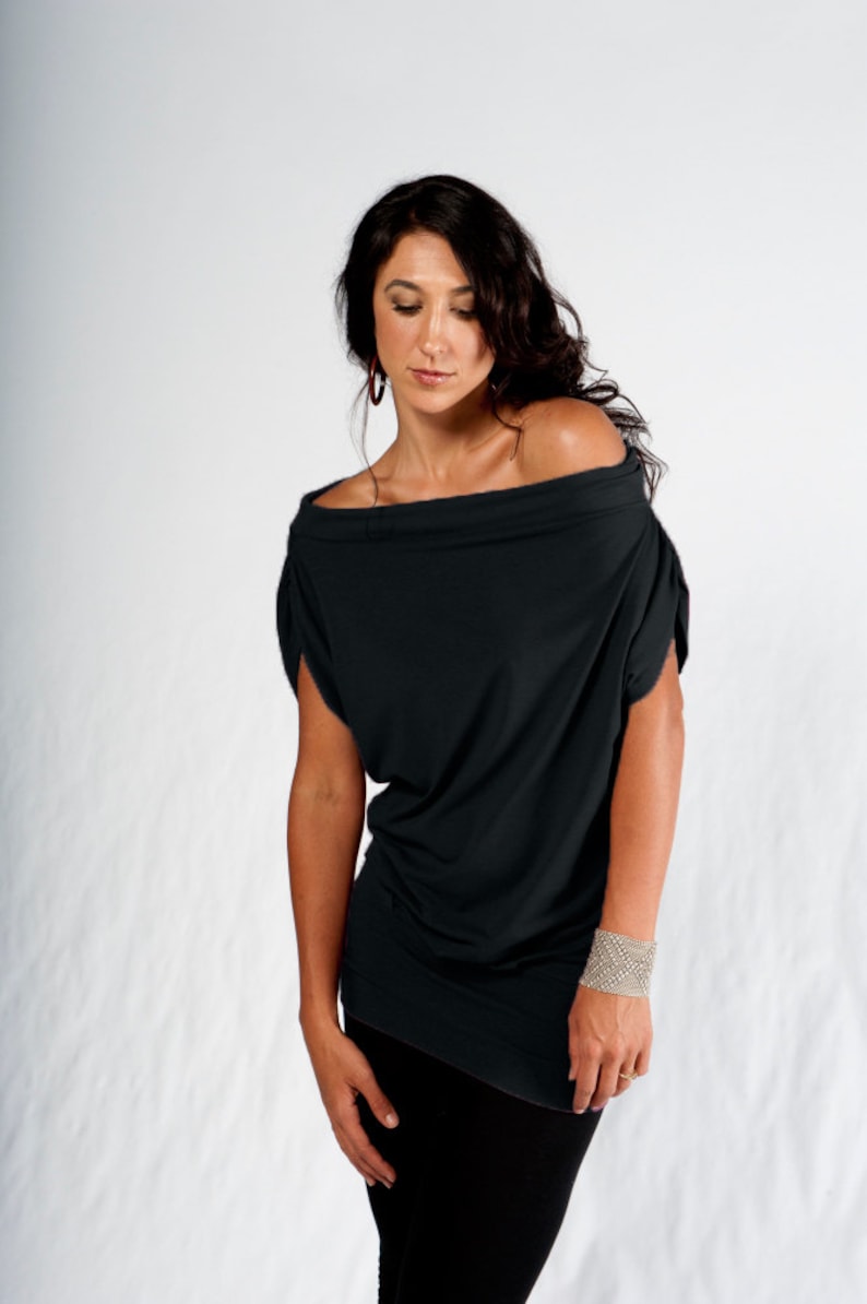 May include: A woman wearing a black off-the-shoulder top with a loose fit and long sleeves. She is wearing black pants and a silver bracelet on her right wrist.
