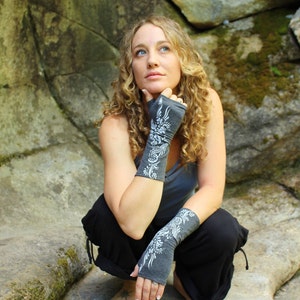 May include: A woman wearing a pair of gray fingerless gloves with a white floral design. The gloves are made of a soft, stretchy fabric and are perfect for keeping your hands warm while still allowing you to use your fingers.