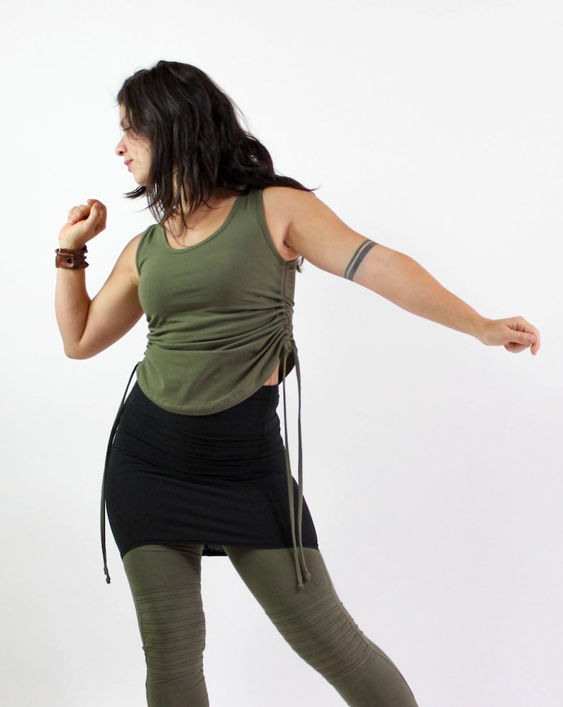 May include: Olive green tank top with drawstring detail, paired with a black mini skirt and olive green leggings. The top has a scoop neckline and a fitted silhouette. The leggings have a ribbed texture. The outfit is casual and comfortable.