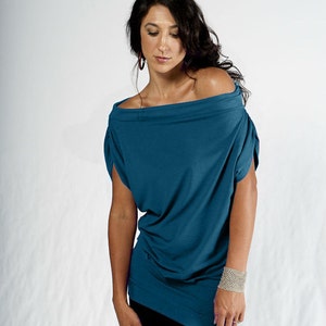 May include: A woman wearing a teal blue off-the-shoulder top with a loose fit and short sleeves. She is wearing black pants and a silver bracelet on her wrist.