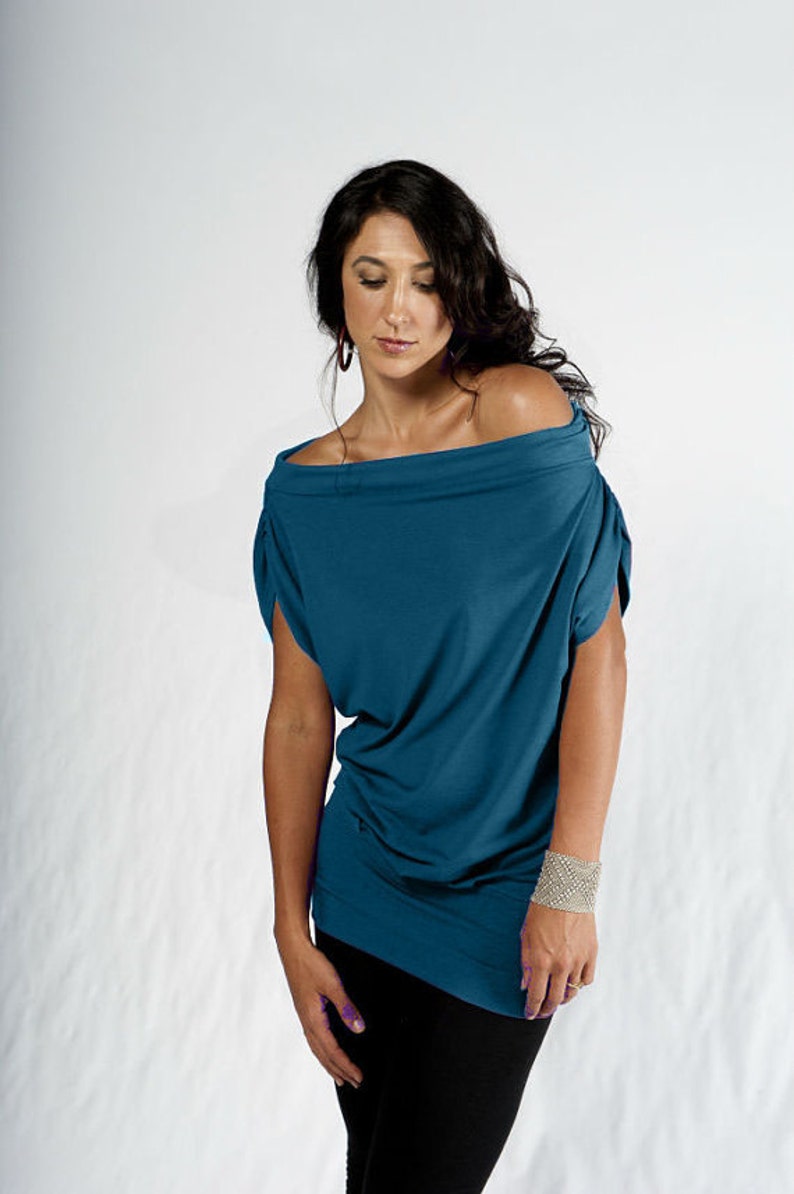 May include: A woman wearing a teal blue off-the-shoulder top with a loose fit and short sleeves. She is wearing black pants and a silver bracelet on her wrist.