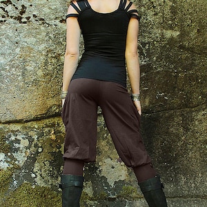 May include: A woman wearing a black top with cut-out shoulder straps and brown pants. She is standing in front of a rocky wall and wearing black boots.