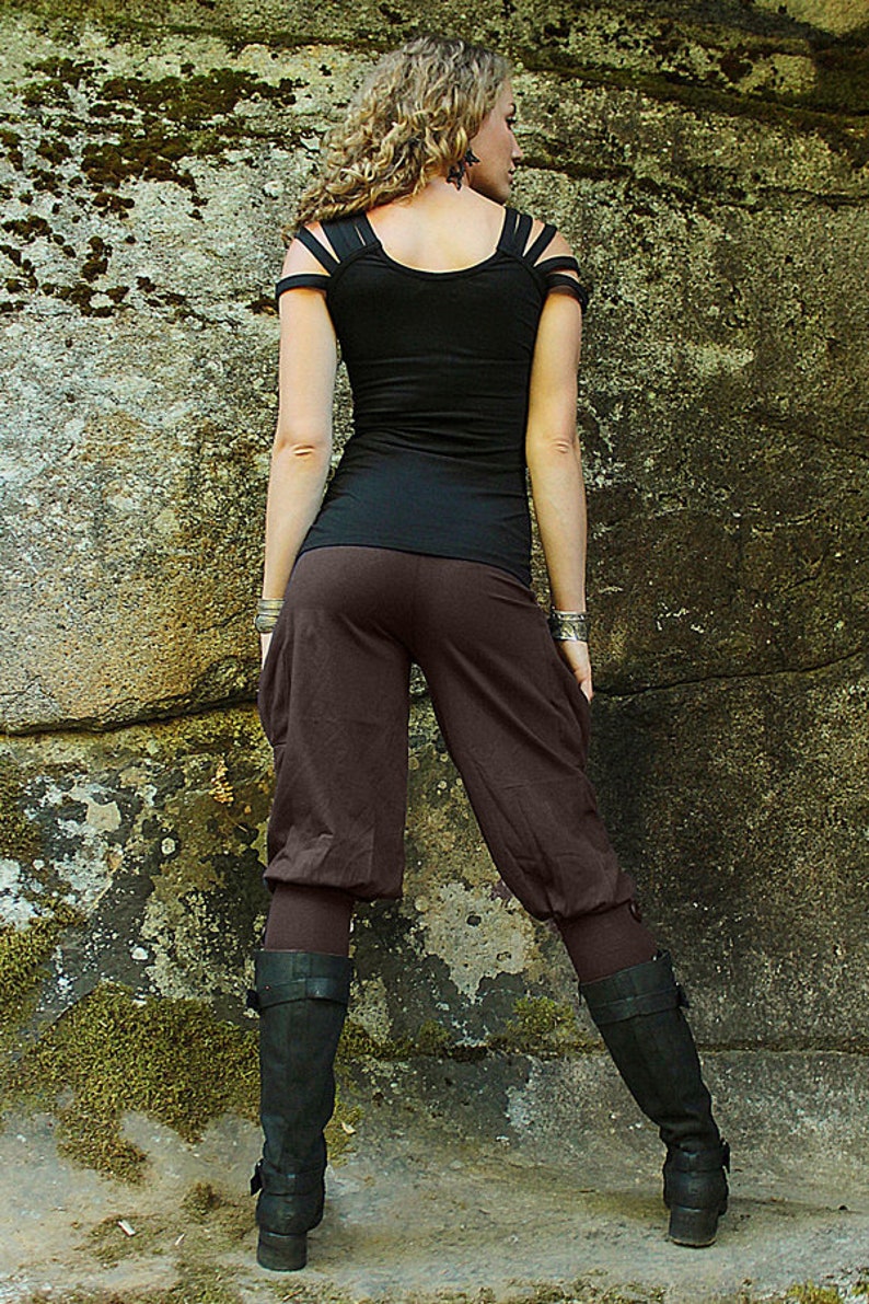 May include: A woman wearing a black top with cut-out shoulder straps and brown pants. She is standing in front of a rocky wall and wearing black boots.