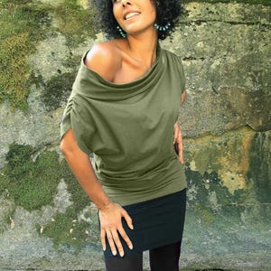 Op de afbeelding: Een vrouw draagt een groen off-shoulder top met een cowl neck en zwarte leggings.