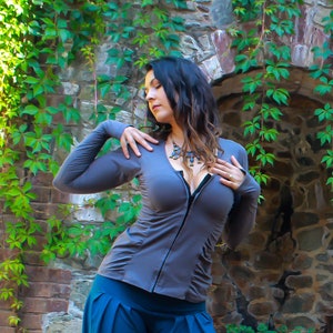 May include: A woman wearing a gray long-sleeved shirt with a zipper in the front. The shirt has a gathered detail at the waist. She is wearing a blue skirt and a silver necklace with a blue stone pendant.
