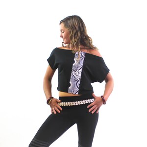 K&ouml;nnte beinhalten: Ein schwarzes Crop-Top mit einem wei&szlig;en Muster und einem schwarzen, schulterfreien Design. Das Top wird mit schwarzen Leggings und einem goldenen Perleng&uuml;rtel kombiniert.