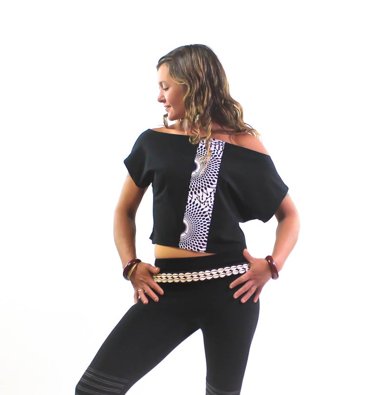 K&ouml;nnte beinhalten: Ein schwarzes Crop-Top mit einem wei&szlig;en Muster und einem schwarzen, schulterfreien Design. Das Top wird mit schwarzen Leggings und einem goldenen Perleng&uuml;rtel kombiniert.