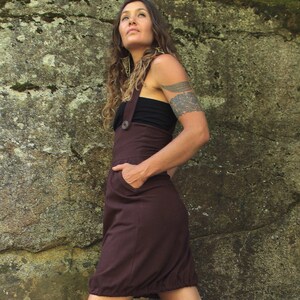 May include: A woman wearing a brown overall dress with a black top underneath. She is standing in front of a rocky wall with moss growing on it.
