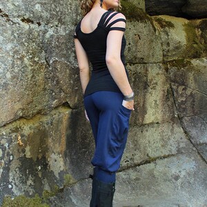 May include: A woman wearing a black top with cut-out shoulders and blue pants. She is standing in front of a rocky wall and wearing black boots.